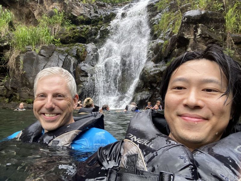 Two men in wetsuits smiling for a selfie in a natural pool at the base of a waterfall, with other swimmers in the background